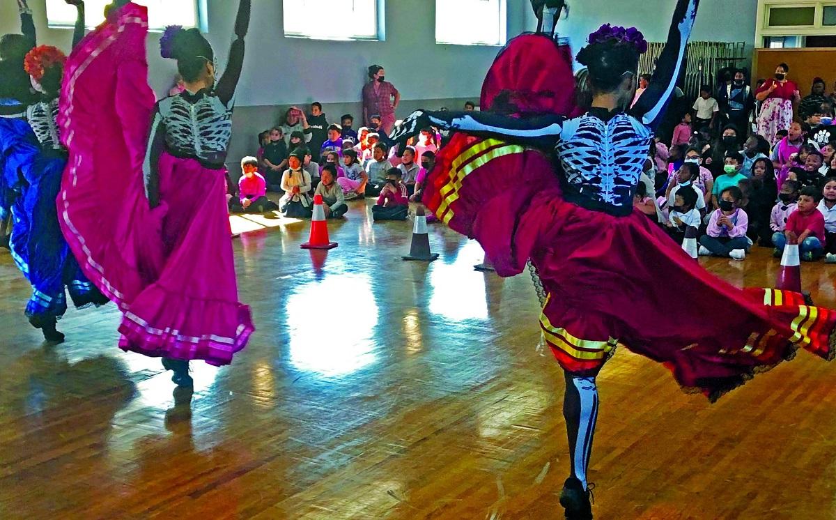 Female dancers, wearing leotards deocrated with a skeleton and colorful skirts, kick their right let in the air.