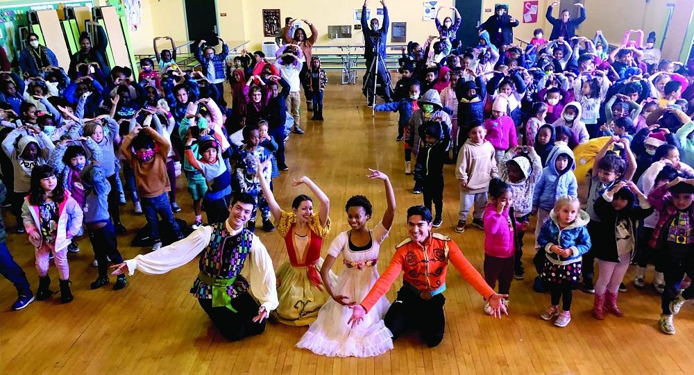 Four performers from OBC's "The Nutcracker" pose in the middle of the aisle while elementary students hold their arms above their head with fingers touching.